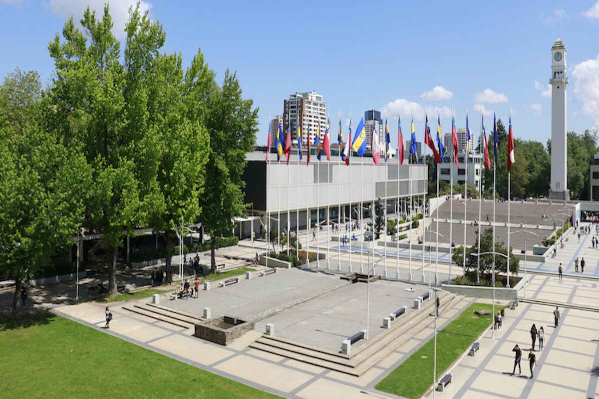 Vista de la plaza del estudiante, con las banderas de Chile y la Universidad puestas. De fondo, la Facultad de Ciencias Químicas en un día soleado.
